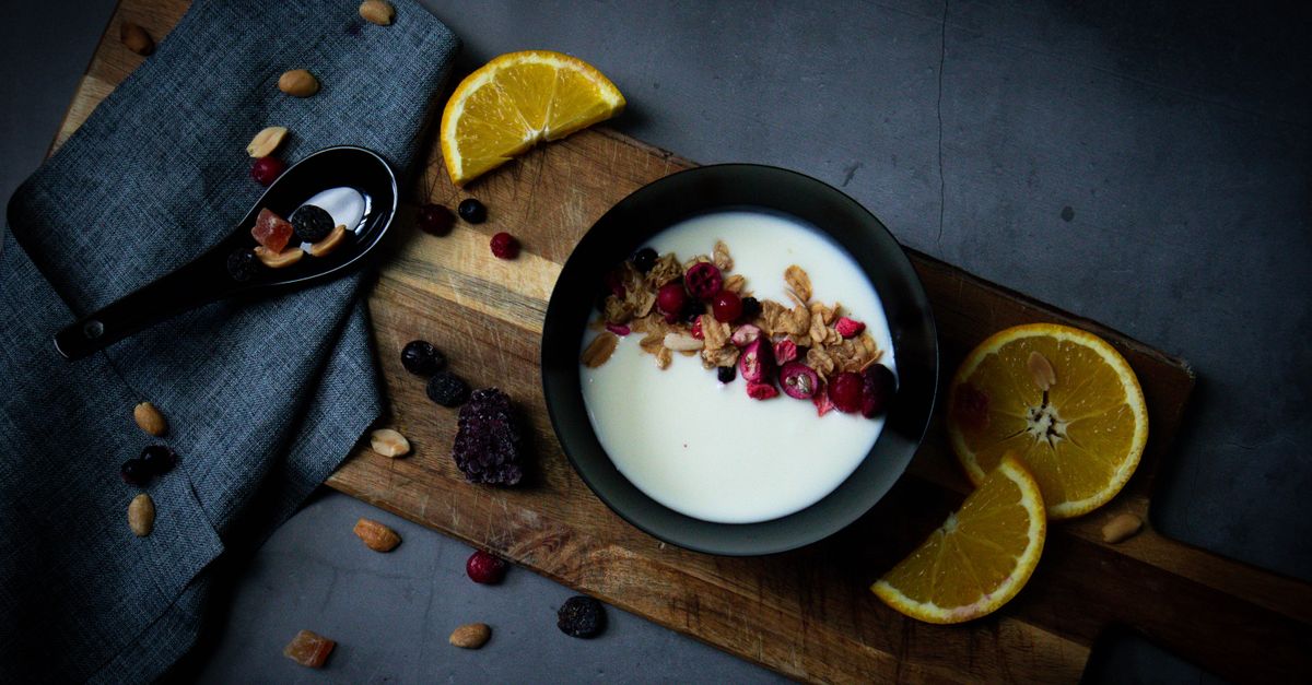 Eine Schüssel Joghurt mit Granola und getrockneten Cranberries steht auf einem Holzbrett, umgeben von Orangenscheiben, Nüssen und getrockneten Beeren.