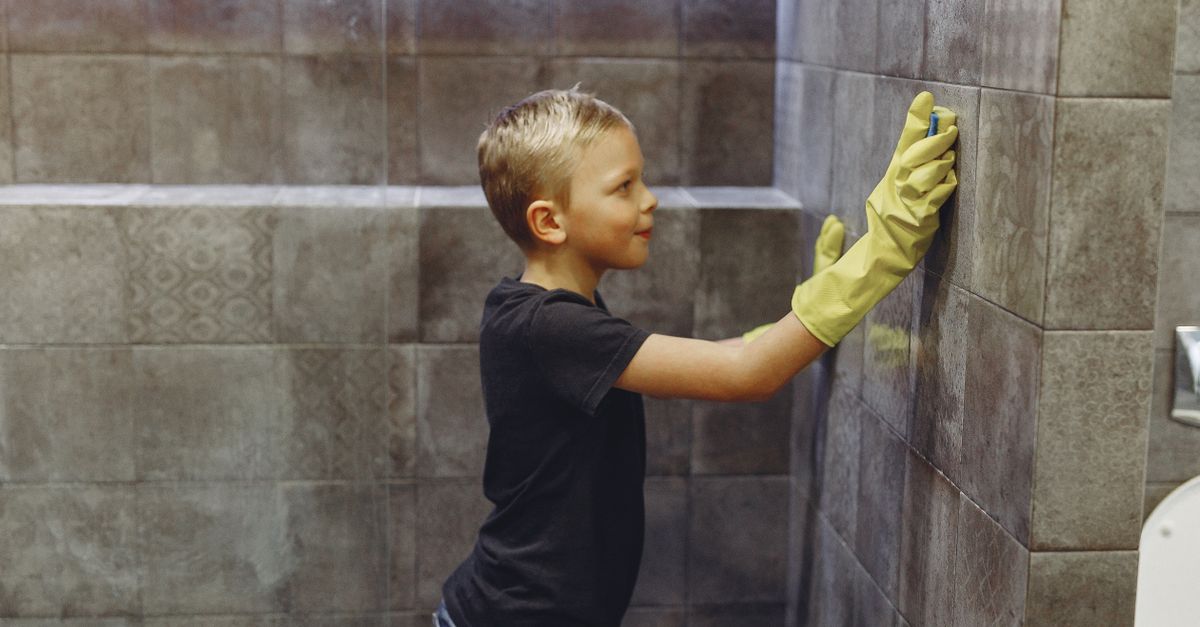 Ein Kind reinigt mit gelben Gummihandschuhen und einem Schwamm die Fliesen einer Dusche.