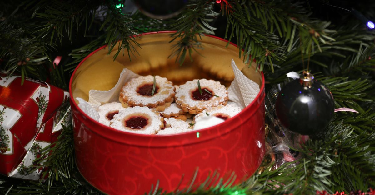 Weihnachtsplätzchen mit Marmeladenfüllung liegen in einer roten Keksdose, umgeben von Tannenzweigen und Weihnachtsschmuck.