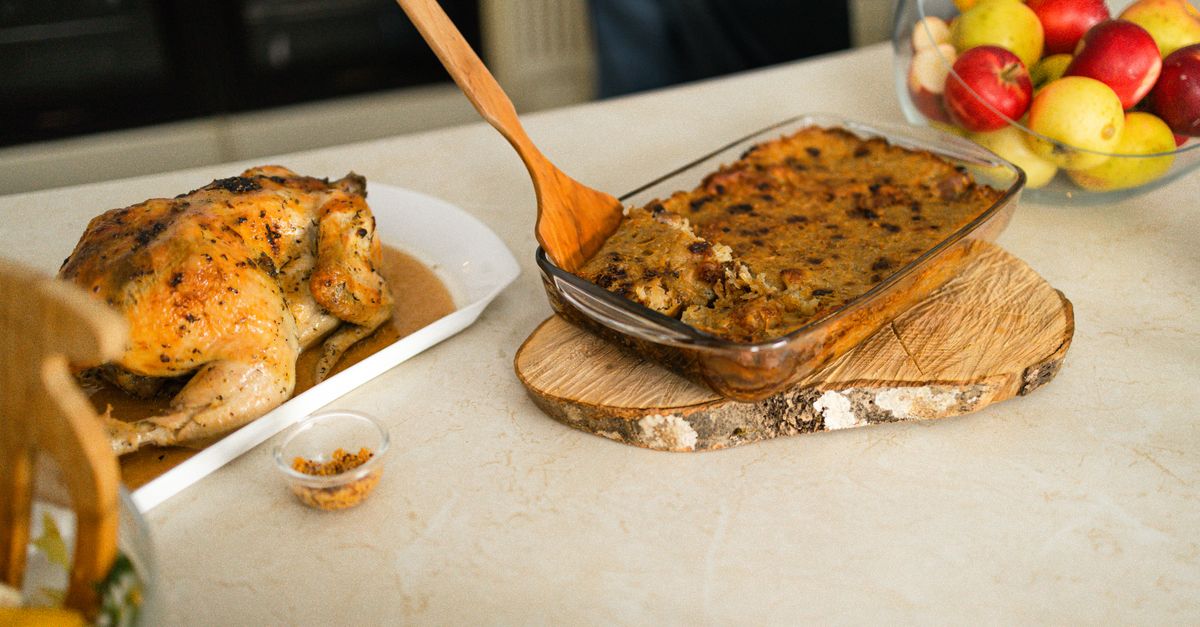 Ein ganzer Brathähnchen liegt neben einer gratinierten Kartoffel-Auflaufform mit goldbrauner Kruste, daneben ein Holzlöffel und eine Schüssel Äpfel.