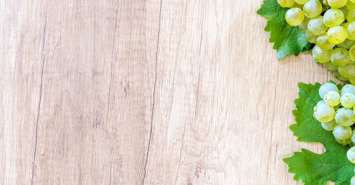 Ein Holztisch mit grünen Weintrauben und Weinblättern am rechten Bildrand, viel freier Raum links.