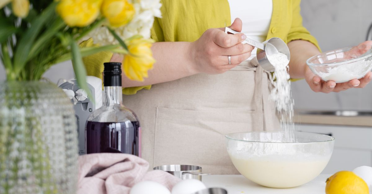 Eine Frau in gelber Bluse gießt Zucker oder Mehl aus einem Messbecher in eine Rührschüssel mit Milch, umgeben von Zitronen, einer Flasche und gelben Tulpen.