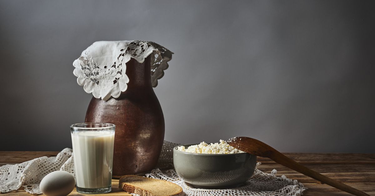 Ein Glas mit Buttermilch oder Milch steht neben einem Tonkrug, einer Schüssel Hüttenkäse und einem Ei auf einem rustikalen Holztisch.