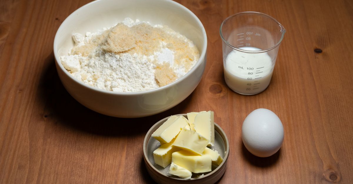 Eine Schüssel mit Mehl, ein Messbecher mit Milch, ein Ei und gewürfelte Butter in einer Holzschale auf einem Holztisch.